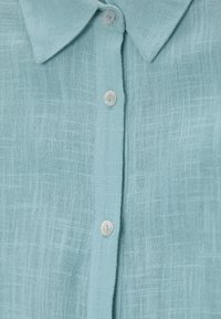 Chemise bleu clair à manches longues en tissu texturé, dotée d'un col et de cinq boutons irisés le long de la patte de boutonnage.