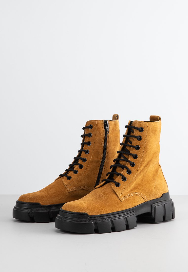 Botas de tobillo de ante en color mostaza con cordones negros y cremallera lateral. Suela de goma negra gruesa, textura suave y puntera reforzada.