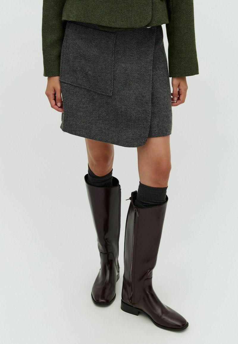 Jupe enveloppante en chevrons gris avec une poche avant, associée à des bottes bordeaux brillantes jusqu'aux genoux et des chaussettes noires.