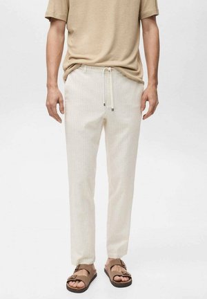 Homme portant une chemise beige à manches courtes, un pantalon crème clair à cordon de serrage et des sandales marron à double bride, debout devant un fond uni.