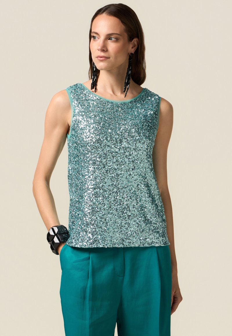 Glittrande turkos paljettopp med rundad halsringning och ärmlös design. Matchas med livfulla turkosa byxor och ett svart geometriskt armband.