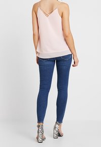 Femme portant un haut sans manches rose pâle avec des bretelles croisées dans le dos, un jean skinny bleu et des sandales à talons hauts au motif python.