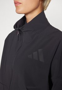 Black athletic jacket featuring a high collar, zip closure, and a geometric logo design on the left side. Smooth, lightweight material.