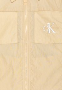 Ljus beige jacka med dragkedja fram, två bröstfickor och vit broderad logotyp. Slätt, lättviktigt tyg med subtil textur.