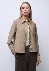 Žena v svetle hnědé košili s lemováním v leopardím vzoru přes béžový top a tmavě hnědé kalhoty, stojící proti jednoduchému světlému pozadí.