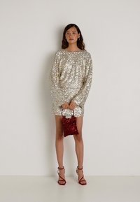 Srebrna mini obleka s svetlečimi pajkicami in dolgimi rokavi, v kombinaciji z majhno rdečo torbico s svetlečimi pajkicami in rdečimi vezalkami, na enobarvni podlagi.