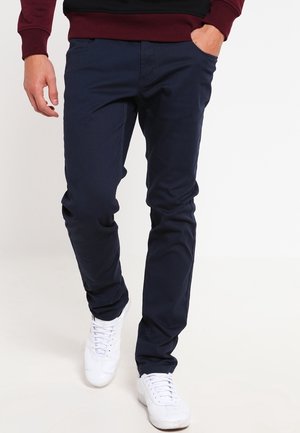 Pantaloni in cotone blu navy con vestibilità slim, dotati di tasche laterali e una texture liscia, abbinati a sneaker bianche.