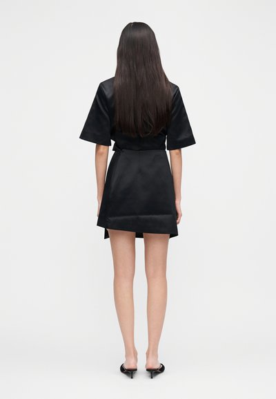 Kvinne med langt, rett hår iført en kort svart kjole og svarte mules med hæl, stående med ryggen vendt mot en ensfarget hvit bakgrunn.