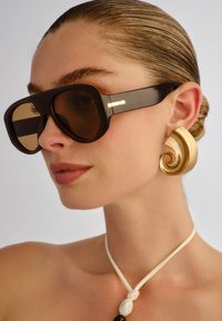 Γυναίκα με πίσω χτενισμένα μαλλιά, φορά μεγάλα μαύρα γυαλιά ηλίου, χρυσό σπείρα σκουλαρίκι και λευκό κορδόνι λαιμού με χάντρα.