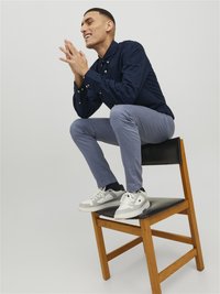 Marineblaues Hemd mit Knöpfen, hellgraue Chinos und grau-weiße Turnschuhe. Person sitzt auf einem Holzstuhl mit schwarzem Sitz.