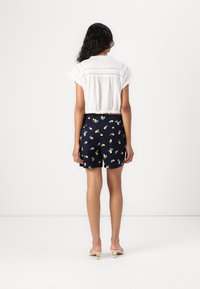 Blusa blanca de corte recortado con detalle de encaje, combinada con pantalones cortos azul marino con un patrón floral. Lleva sandalias beige. Se muestra la vista trasera.