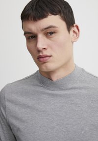 Camisa gris de manga larga con un cuello redondeado alto. Textura suave y lisa con un mínimo detalle de costura y corte recto.