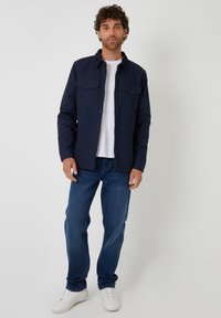 Granatowa koszula z guzikami i dwoma kieszeniami na piersi, zestawiona z dżinsami i białymi trampkami. Koszula ma klasyczny kołnierzyk oraz teksturowany materiał.