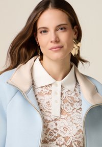 Žena s dlouhými hnědými vlasy nosící velké zlaté geometrické náušnice, modrou bundu s béžovým límcem a bílý krajkový top s límečkem, neutrální pozadí.