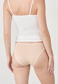 Žena zezadu v bílém bezramínkovém topu a béžovém bezešvém spodním prádle na neutrálním pozadí.