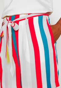Shorts rayés présentant des rayures verticales rouges, blanches et turquoise, fabriqués dans un tissu léger avec un détail de ceinture nouée.