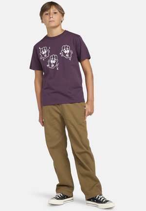 Fioletowa bawełniana koszulka z białym nadrukiem rąk z literami. Połączona z khaki spodniami i czarnymi trampkami z białymi podeszwami.