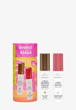 Set aus drei kosmetischen Produkten in einer zylindrischen Box. Die Box zeigt ein leuchtend pinkes und gelbes Design mit tropischen Mustern. Zwei Flaschen sind weiß mit farbigen Deckeln – einer braun und einer pink.