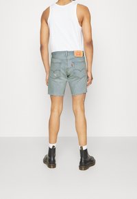 Man wearing light blue denim shorts with frayed hems, white tank top, white socks, and black lace-up boots, standing against a plain wall.