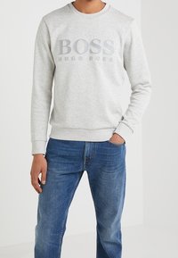 Mann in hellgrauem Sweatshirt mit dem Schriftzug "BOSS HUGO BOSS" und blauen Jeans, steht vor einem schlichten weißen Hintergrund.