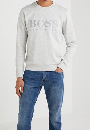 Hombre vestido con un sudadera gris claro con el texto "BOSS HUGO BOSS" y jeans azules, de pie contra un fondo blanco liso.