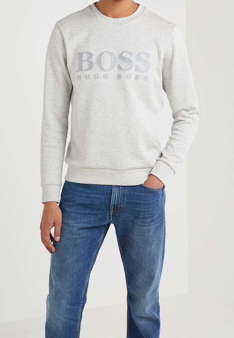 Hombre vestido con un sudadera gris claro con el texto "BOSS HUGO BOSS" y jeans azules, de pie contra un fondo blanco liso.