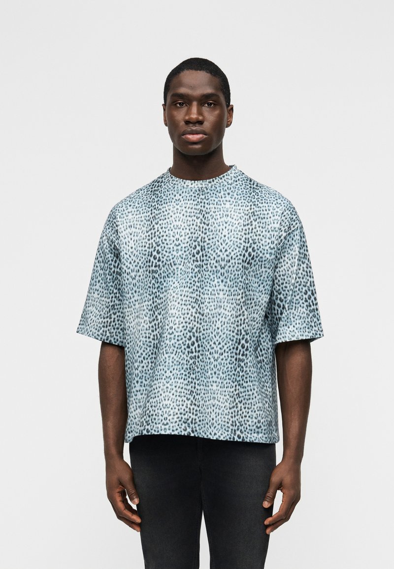 Άνδρας που φοράει φαρδιά μπλε και λευκή πουκάμισα με κοντά μανίκια και σχέδιο λεοπάρδαλης, καθώς και μαύρο παντελόνι, στέκεται μπροστά σε λευκό απλό φόντο.