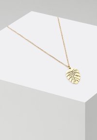 Collana con pendente a forma di foglia color oro, caratterizzata da un design ritagliato dettagliato, appesa a una delicata catena su un supporto espositivo geometrico.