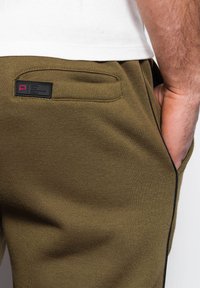 Olivgrüne Jogginghosen mit glatter Textur, ausgestattet mit einer Gesäßtasche und einem Markenetikett, verziert mit schwarzem Piping entlang der Kanten.