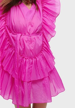 Vrouw in een felroze jurk met ruches, een riem en een dikke ketting, tegen een effen lichte achtergrond.