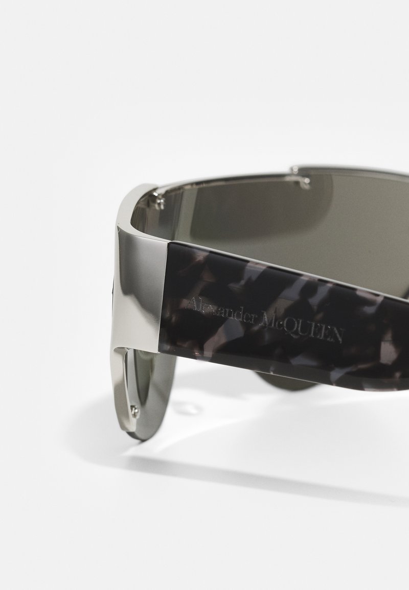 Metall-Sonnenbrille mit dunkelgrauen Gläsern, ausgestattet mit einem glänzenden Schildpatt-Bügel, der durch eine silberne Logo-Plakette akzentuiert wird. Schlankes und modernes Design.