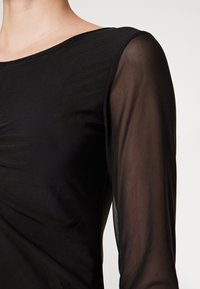 Czarna bluzka z długimi rękawami o półprzezroczystej fakturze. Okrągły dekolt i gładki materiał z delikatnym marszczeniem z przodu. Eleganckie detale w designie.