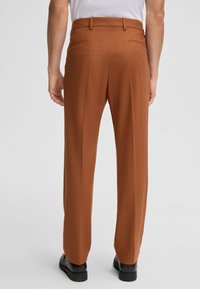 Bruine op maat gemaakte broek van een textuurstof, met plooien aan de voorkant, twee achterzakken en een recht model. Gemaakt met zwarte schoenen.