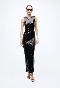 Black sequined gown with a fitted silhouette, cap sleeves, and a floor-length hem. Model wearing black sunglasses and strappy sandals.
