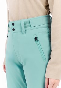 Close-up van teal waterdichte broek met verstelbare tailleband, ritssluiting zak en een beige textuur lange mouwen top aan de rechterzijde van de drager.