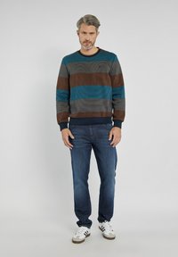 Pasiasty sweter w ciemnoniebieskim, brązowym i teal, z prążkowanym dekoltem i mankietami. Połączony z dopasowanymi niebieskimi dżinsami i białymi butami sportowymi.
