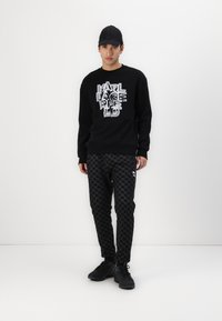 Svart sweatshirt med en texturerad logotyp i vitt, ihop med svarta mönstrade byxor i hundtandsmönster och en liten logotypdetalj.