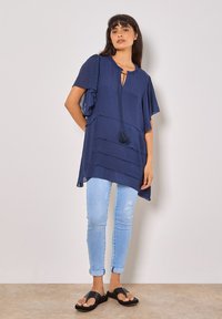 Marineblaues Tunika mit kurzen, rüschierten Ärmeln, einem Schlüsselloch-Ausschnitt mit Quaste, gestuftem Design, kombiniert mit hellblauen Skinny Jeans und schwarzen Sandalen.