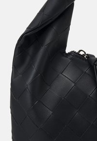 Sac en cuir noir avec un motif carré tissé et une tirette de fermeture éclair argentée visible sur un fond clair uni.