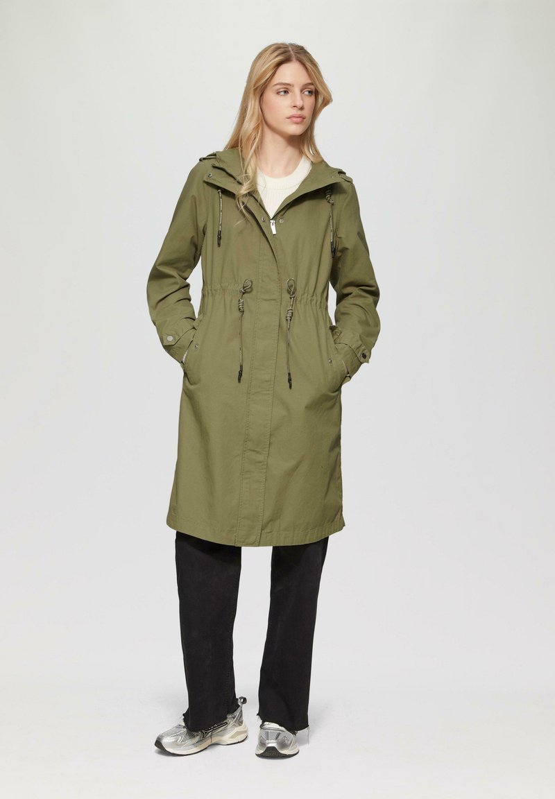 Oliwkowa, średniej długości kurtka z regulowanym pasem, odpinanym kapturem i przednimi kieszeniami. Nosi ją osoba w czarnych spodniach i srebrnych trampkach.