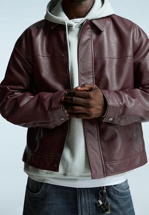 Persoon draagt een donkerrode leren jas over een lichtgrijze hoodie met gevouwen handen en een spijkerbroek met een sleutelhanger bevestigd aan een broekriemlus.