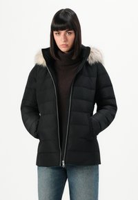 Giacca a vento nera con zip frontale, dotata di cappuccio foderato in pelliccia, design trapuntato e tasche laterali, abbinata a un maglione dolcevita marrone.