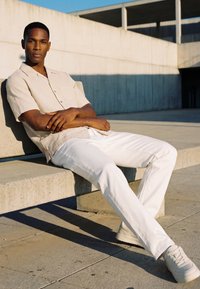 Ein Mann trägt ein hellbeiges kurzärmeliges Hemd und weiße Hose und sitzt auf einer Betonbank im hellen Sonnenlicht vor einem modernen Betongebäude.