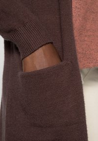 Brun strikket cardigan med ribbede manchetter og en sidelomme. Blød tekstur med et lagdelt udseende mod en lys baggrund.