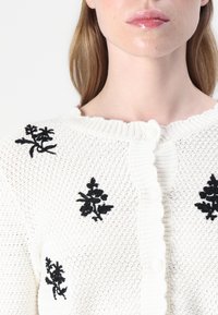 Cardigan bianco lavorato a maglia con ricami floreali neri. Presenta un colletto scalloppato e chiusura a bottone, motivo a maglia testurizzato.