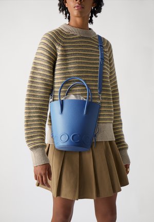 Borsa a secchiello in pelle blu con superficie texturizzata, doppi manici e tracolla regolabile. Indossata con un maglione a righe e una gonna plissé.