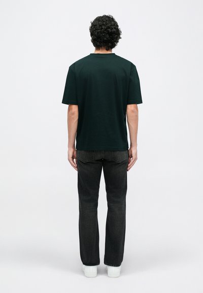 Sötétzöld, rövid ujjú pamut póló, fekete egyenes szárú farmernadrággal és fehér sportcipővel párosítva, hátulról nézve.