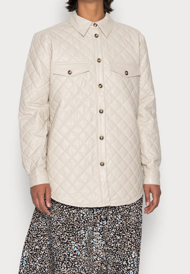 Femme portant une veste crème matelassée à boutons avec poches à rabat, sur une jupe noire avec des motifs floraux blancs et bleus.