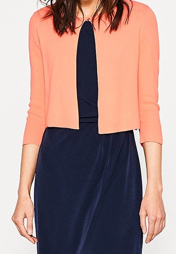 Femme portant une robe bleu marine avec un cardigan court corail ajusté à manches trois-quarts, debout, bras détendus le long du corps.