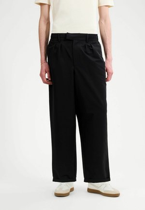 Persona che indossa pantaloni neri larghi con orli risvoltati, abbinati a scarpe da ginnastica bianche e una camicia beige a maniche corte infilata, su sfondo bianco.